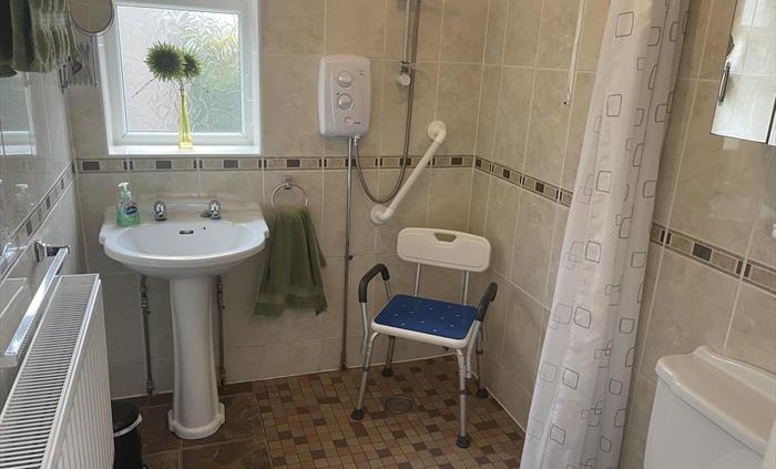 shows sink, window, electric shower, radiator, shaving mirror, electric shaver socket, mirror cupboard, hand grip and a height adjustable shower chair